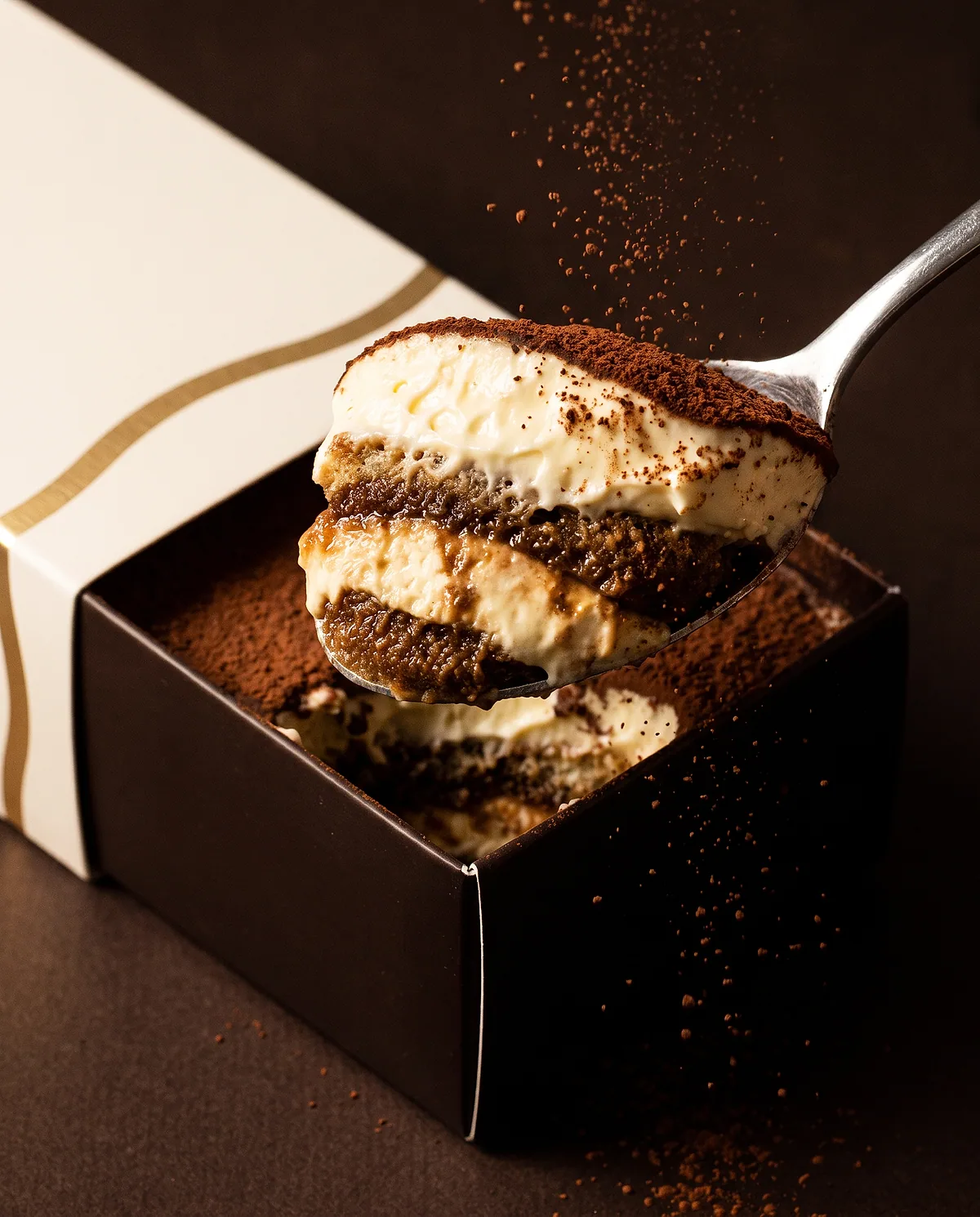 Gros plan d'une cuillère soulevant les couches d'un tiramisu, cacao en suspension.